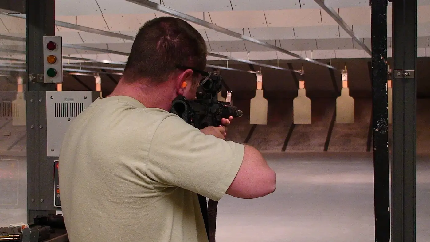 Shooter wearing eye and ear protection aiming a rifle downrange in an indoor shooting lane