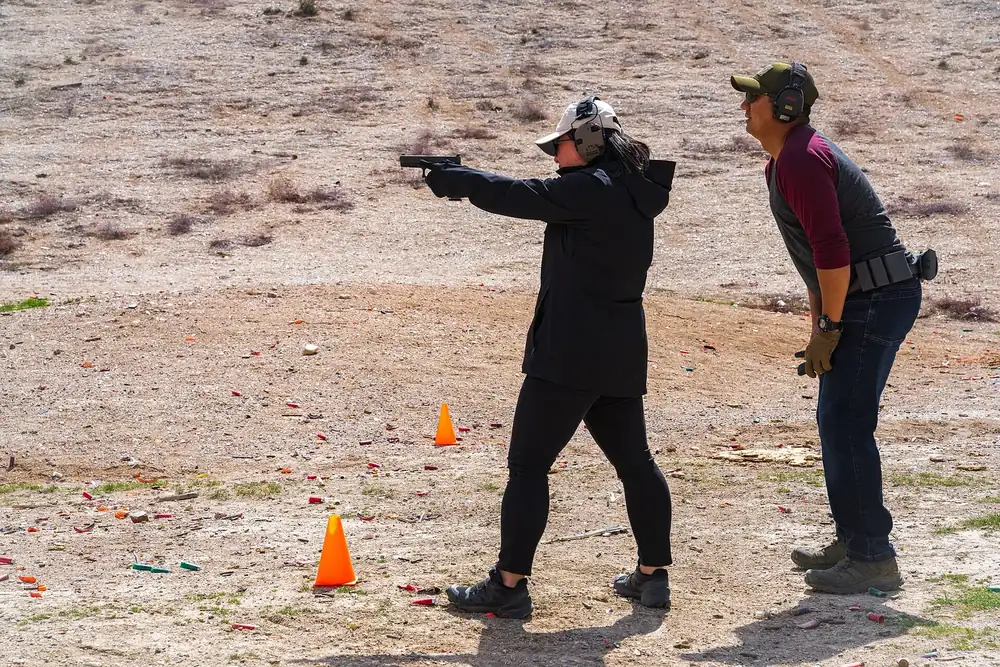 Shooter in earmuffs and safety glasses firing a pistol as an instructor in ear protection watches