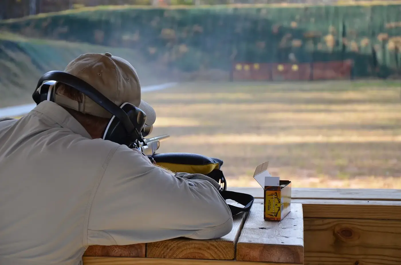 Shooter in earmuffs firing a rifle from a bench rest at an outdoor range